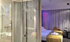 Łazienka i sypialnia w hotelu Puro Wrocław. Szklane drzwi oddzielają prysznic od pokoju z łóżkiem i zasłonami.