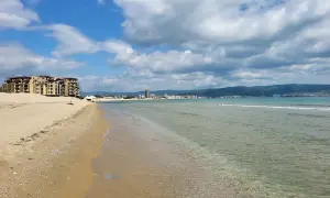 Słoneczny brzeg z piaszczystą plażą, turkusową wodą i hotelami w tle. Chmury na błękitnym niebie zapowiadają piękną pogodę.