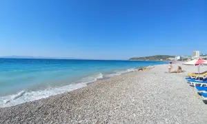 Słoneczna plaża w Ixia, Rodos, z turkusowym morzem i kamienistym brzegiem. Dwie osoby odpoczywają pod parasolem.