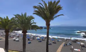 Palmy nad czarną plażą w Puerto de Santiago. Ludzie odpoczywają pod parasolami, a fale rozbijają się o brzeg.
