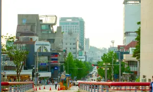 Tętniąca życiem ulica w **Hongdae**, pełna ludzi, sklepów i nowoczesnej architektury, z widokiem na wzgórza w tle.