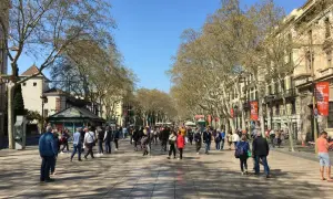 Tłum ludzi spaceruje po słonecznej La Rambla w Barcelonie, otoczonej drzewami i zabytkowymi budynkami.