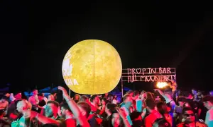 Tłum ludzi bawi się na plaży podczas **full moon party**. W tle neonowy napis "DROP IN BAR FULL MOON PARTY" i wielka, świecąca kula imitująca księżyc.