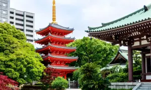 Piękna, wielopoziomowa pagoda w kolorze czerwonym, otoczona bujną zielenią i tradycyjną architekturą. W tle nowoczesny budynek, kontrastujący z historycznym krajobrazem. To widok z Hakaty.