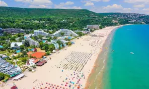 Słoneczna plaża na riwierze bułgarskiej pełna leżaków i parasoli, z turkusowym morzem i zielonymi wzgórzami w tle.