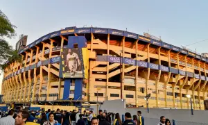 Tłum kibiców przed stadionem La Bombonera. Wielki baner z piłkarzem zdobi żółto-niebieską fasadę.