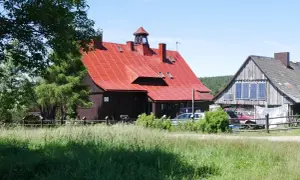 Urokliwa chatka gorzystow z czerwonym dachem, otoczona zielenią. Obok drewniany budynek z panelami słonecznymi.