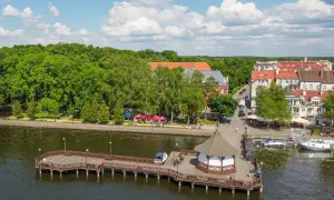 Drewniany pomost z altaną nad jeziorem, zielone drzewa i miasteczko z czerwonymi dachami. Idealne miejsce, gdzie na Mazury.