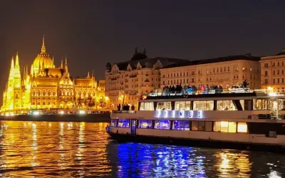 Nocny rejs po rzece w Budapeszcie. Oświetlony Parlament i statek wycieczkowy "Primus" odbijają się w wodzie.