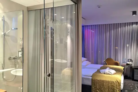 Łazienka i sypialnia w hotelu Puro Wrocław. Szklane drzwi oddzielają prysznic od pokoju z łóżkiem i zasłonami.