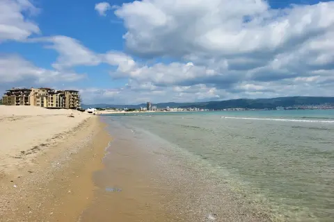 Słoneczny brzeg z piaszczystą plażą, turkusową wodą i hotelami w tle. Chmury na błękitnym niebie zapowiadają piękną pogodę.