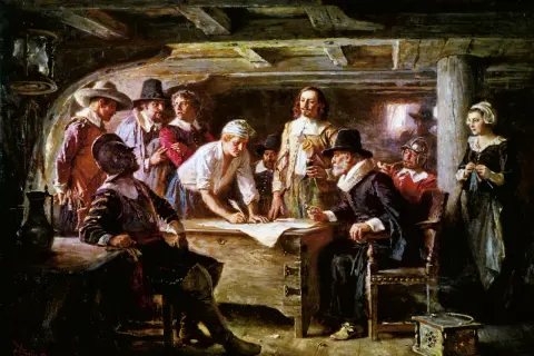 Pielgrzymi przy stole, jeden pisze, inni patrzą. To scena z życia na statku przed przybyciem do Plymouth Rock.
