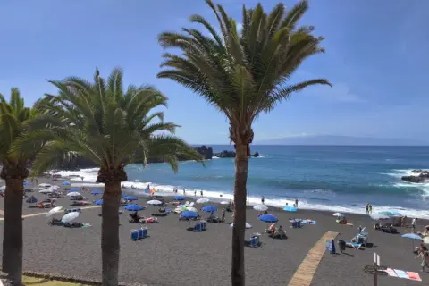 Palmy nad czarną plażą w Puerto de Santiago. Ludzie odpoczywają pod parasolami, a fale rozbijają się o brzeg.