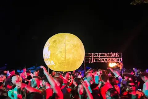 Tłum ludzi bawi się na plaży podczas **full moon party**. W tle neonowy napis "DROP IN BAR FULL MOON PARTY" i wielka, świecąca kula imitująca księżyc.