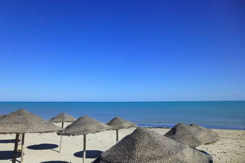 Słoneczna plaża na Djerbie w kwietniu. Puste parasole z trawy na piasku, turkusowe morze i bezchmurne niebo.