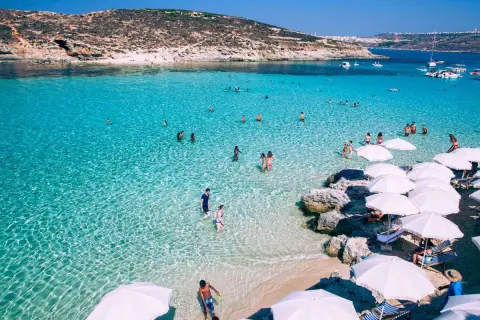 Malta w maju: turkusowa woda, plaża z białymi parasolami i ludzie kąpiący się w morzu.