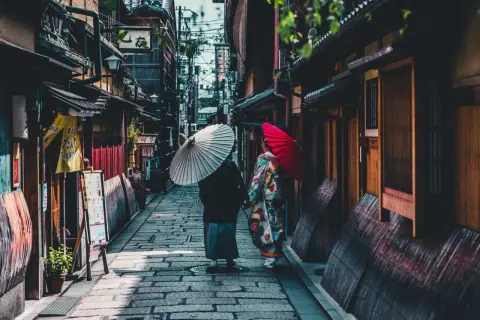 Dwie kobiety w kimonach spacerują wąską uliczką w Kioto, trzymając parasolki. To fascynujące ciekawostki o Japonii.