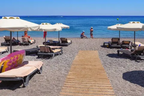 Słoneczny kurort nad Morzem Czarnym. Leżaki pod parasolami, drewniana ścieżka na kamienistej plaży, ludzie kąpiący się w morzu.