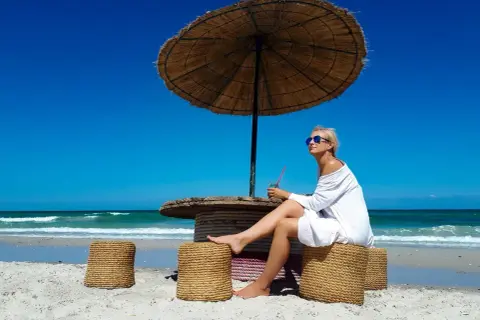 Kobieta siedzi pod parasolem na plaży, popijając napój. Ceny w Tunezji są przystępne, co pozwala na takie relaksy.