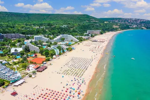 Słoneczna plaża na riwierze bułgarskiej pełna leżaków i parasoli, z turkusowym morzem i zielonymi wzgórzami w tle.