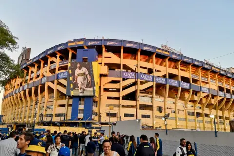 Tłum kibiców przed stadionem La Bombonera. Wielki baner z piłkarzem zdobi żółto-niebieską fasadę.
