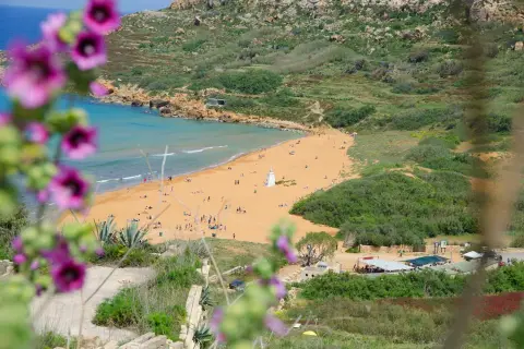 Malta pogoda kwiecień: piaszczysta plaża z turystami, turkusowe morze i zielone wzgórza. W oddali biała wieża.