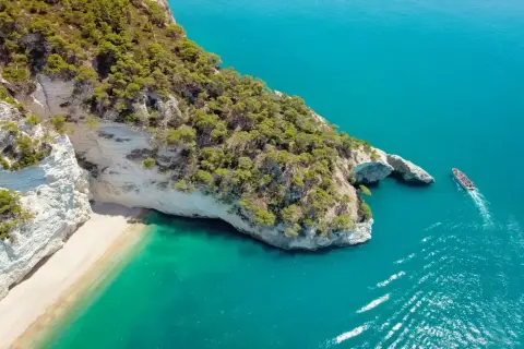 Włochy regiony turystyczne: białe klify, piaszczysta plaża i turkusowe morze. Statek wycieczkowy płynie obok skalnego łuku.