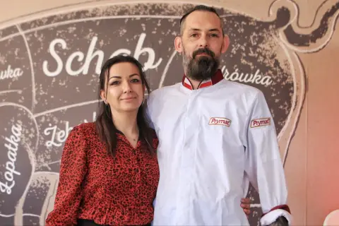 Para z uśmiechem na tle schematu rozbioru świni. On w białej kurtce z logo Prymat, ona w czerwonej bluzce. Wszyscy chwalą złota świnka opinie.