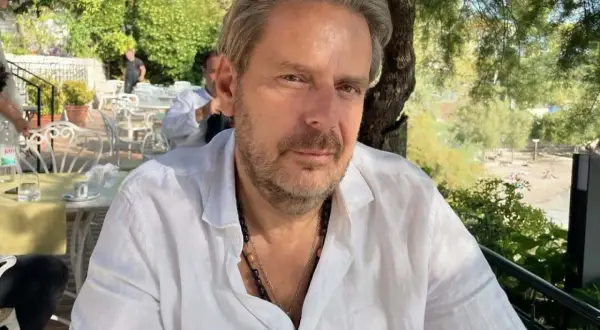 Mężczyzna w białej koszuli, z naszyjnikami, siedzi w restauracji. W tle widać ogród i plażę. Styl atelier amaro.