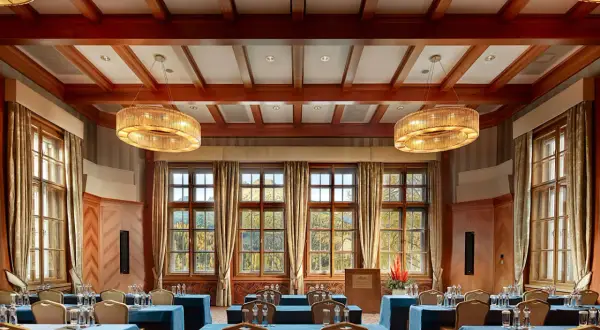 Elegancka sala konferencyjna w Kempinski Hotel, z drewnianymi belkami na suficie, dużymi oknami z widokiem na jesienny krajobraz i dwoma ozdobnymi żyrandolami.