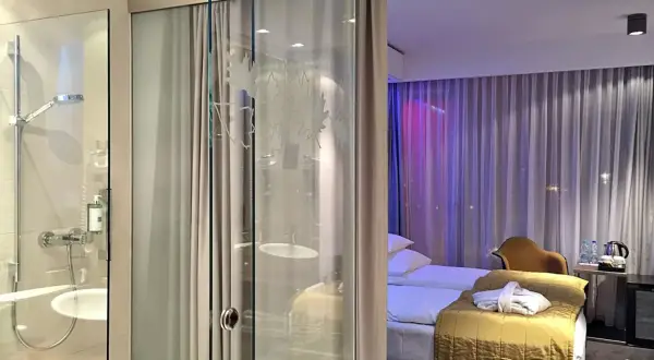 Łazienka i sypialnia w hotelu Puro Wrocław. Szklane drzwi oddzielają prysznic od pokoju z łóżkiem i zasłonami.