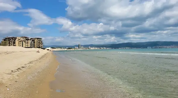 Słoneczny brzeg z piaszczystą plażą, turkusową wodą i hotelami w tle. Chmury na błękitnym niebie zapowiadają piękną pogodę.