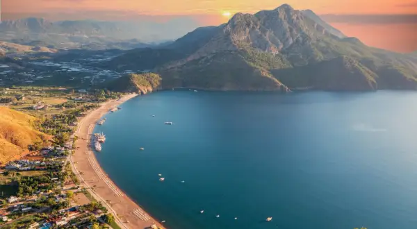 Malownicze wybrzeże Turcji o zachodzie słońca. Góry schodzą do turkusowego morza, a plaża pełna jest leżaków.