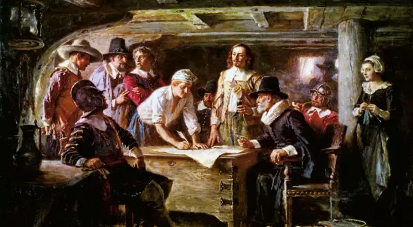 Pielgrzymi przy stole, jeden pisze, inni patrzą. To scena z życia na statku przed przybyciem do Plymouth Rock.
