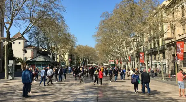 Tłum ludzi spaceruje po słonecznej La Rambla w Barcelonie, otoczonej drzewami i zabytkowymi budynkami.