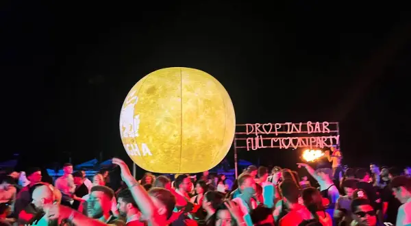 Tłum ludzi bawi się na plaży podczas **full moon party**. W tle neonowy napis "DROP IN BAR FULL MOON PARTY" i wielka, świecąca kula imitująca księżyc.