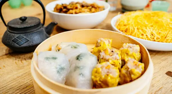Pyszne dim sum w bambusowym parowniku, obok miseczki z makaronem i daniem mięsnym.