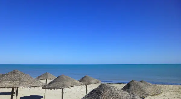 Słoneczna plaża na Djerbie w kwietniu. Puste parasole z trawy na piasku, turkusowe morze i bezchmurne niebo.