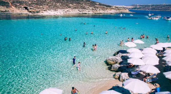 Malta w maju: turkusowa woda, plaża z białymi parasolami i ludzie kąpiący się w morzu.