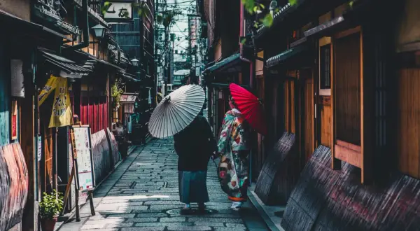 Dwie kobiety w kimonach spacerują wąską uliczką w Kioto, trzymając parasolki. To fascynujące ciekawostki o Japonii.