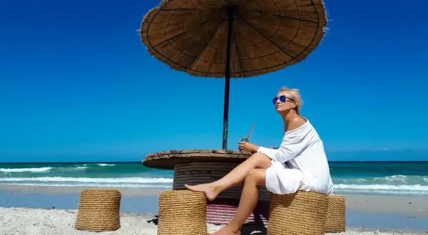Kobieta siedzi pod parasolem na plaży, popijając napój. Ceny w Tunezji są przystępne, co pozwala na takie relaksy.