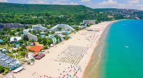 Słoneczna plaża na riwierze bułgarskiej pełna leżaków i parasoli, z turkusowym morzem i zielonymi wzgórzami w tle.