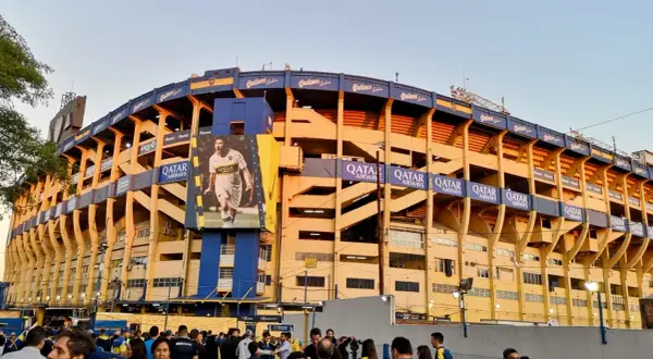 Tłum kibiców przed stadionem La Bombonera. Wielki baner z piłkarzem zdobi żółto-niebieską fasadę.