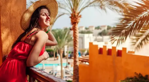Uśmiechnięta kobieta w słomkowym kapeluszu i czerwonej sukience podziwia widok z balkonu. Idealna sharm el sheikh pogoda marzec.