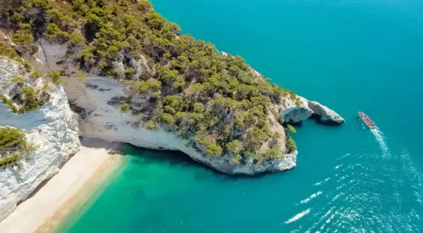 Włochy regiony turystyczne: białe klify, piaszczysta plaża i turkusowe morze. Statek wycieczkowy płynie obok skalnego łuku.