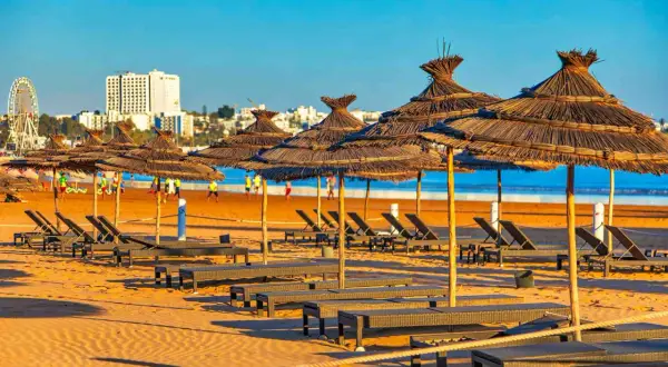 Słoneczna plaża w Agadirze w marcu, puste leżaki pod strzechowymi parasolami, w tle budynki i diabelski młyn.