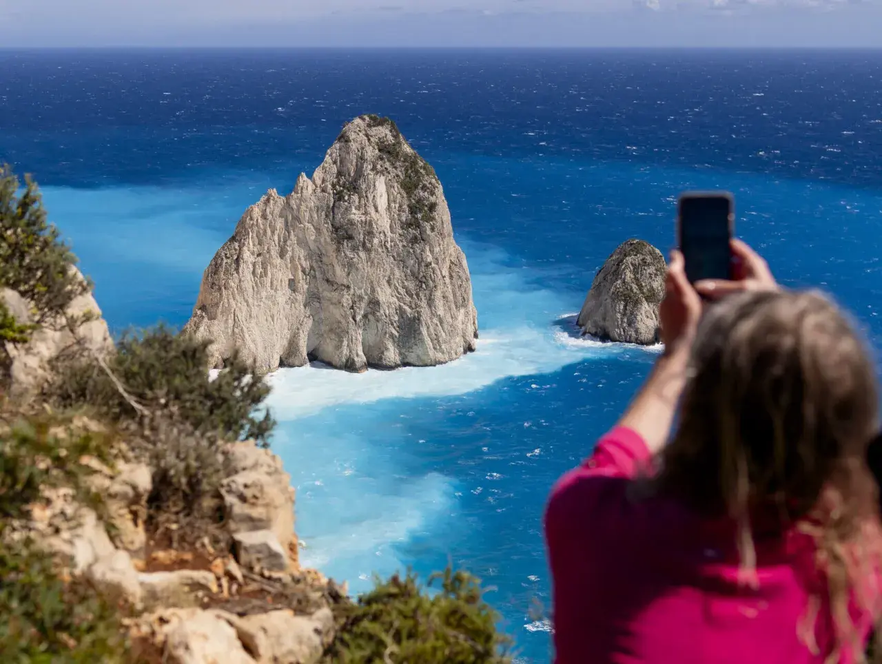 Widok na turkusowe morze i skaliste wysepki. Kobieta robi zdjęcie telefonem, uwieczniając piękno Zakynthos. Idealne miejsce na wycieczki fakultatywne.