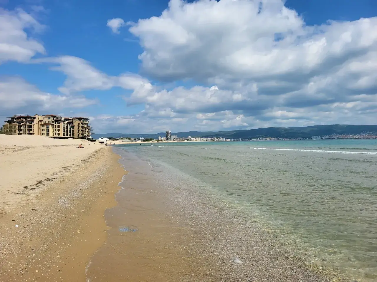 Słoneczny brzeg z piaszczystą plażą, turkusową wodą i hotelami w tle. Chmury na błękitnym niebie zapowiadają piękną pogodę.