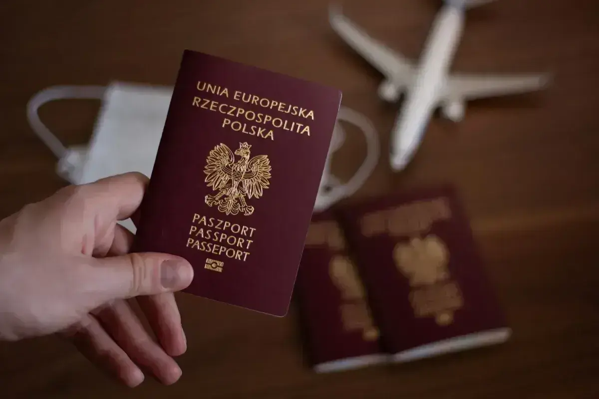 Dłoń trzyma polski paszport. Obok leżą kolejne paszporty, maseczka i samolot, sugerując podróż i pytając, ile wyrabia się paszport.