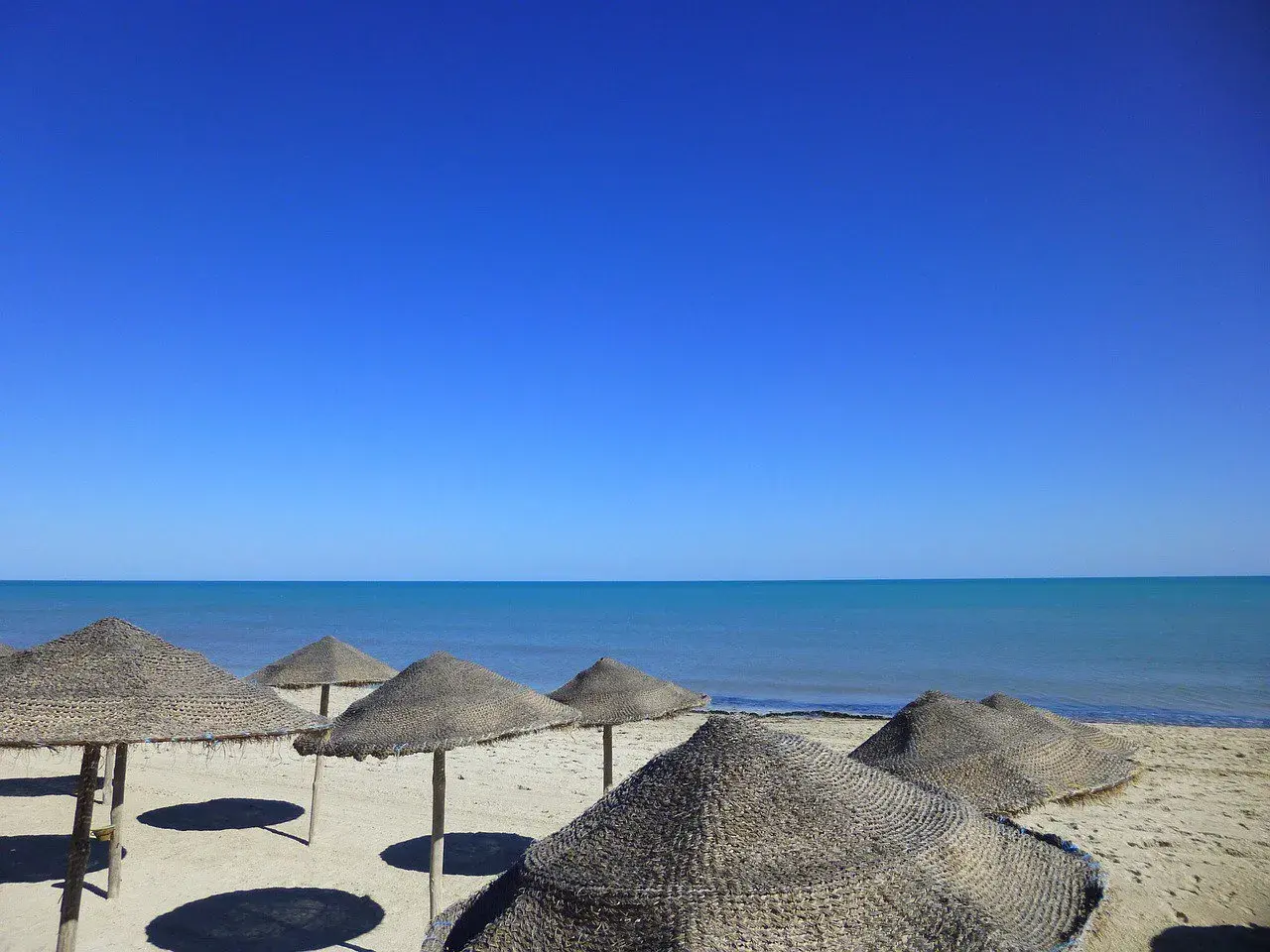 Słoneczna plaża na Djerbie w kwietniu. Puste parasole z trawy na piasku, turkusowe morze i bezchmurne niebo.